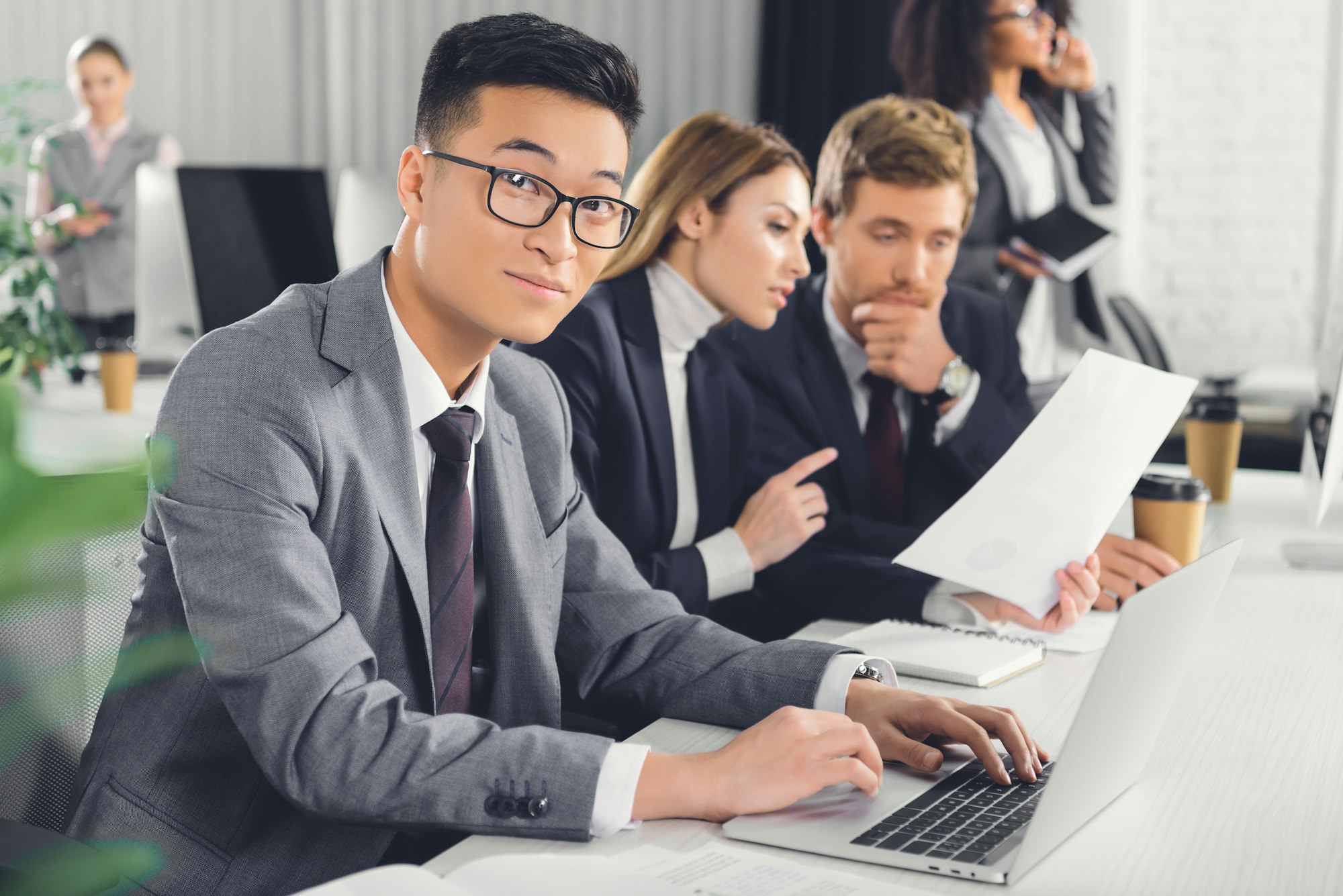 businesspeople working with papers, asian businessman using laptop and looking at camera in office